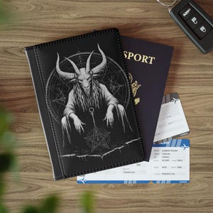 May include: Black passport holder featuring a detailed illustration of a goat-like figure with large horns, a pentagram, and a circular design. A dark blue passport and two boarding passes are next to the holder. A car key is also visible.