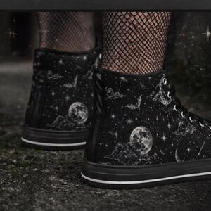 May include: Black high-top sneakers with a celestial design. The sneakers feature a black base with a pattern of moons, stars, and bats. The soles are black with a white stripe.