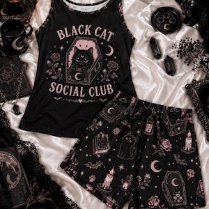 May include: A black tank top and shorts pajama set featuring a pink graphic print. The tank top displays a cat inside a coffin with the text "BLACK CAT SOCIAL CLUB". The shorts feature a repeating pattern of cats, coffins, candles, and roses.
