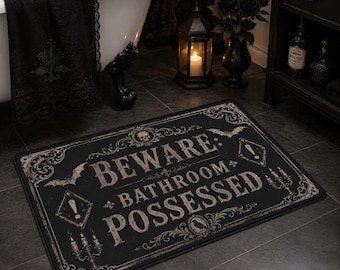 May include: A rectangular black bath mat featuring a beige vintage-style border with ornate scrollwork, bats, a skull, and candelabras. The text in the center reads BEWARE: BATHROOM POSSESSED in a distressed, serif font with exclamation points.