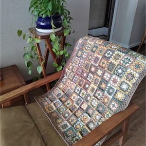 Puede incluir: Una manta de ganchillo con un patrón cuadrado multicolor en tonos marrones, verdes, amarillos y blancos. La manta está drapeada sobre un sillón de madera.