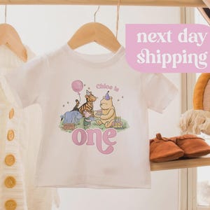 May include: A wooden clothing rack displays a white baby tee with a Winnie the Pooh graphic, a cream-colored cardigan, a rainbow wall hanging, and a tan hat. The tee says "Chloe is one". Brown leather baby shoes are on a shelf.