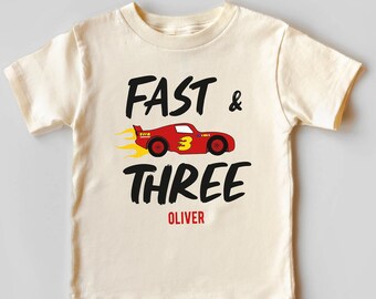 May include: A cream colored t-shirt with a red race car graphic and the text "Fast & Three Oliver" in black.