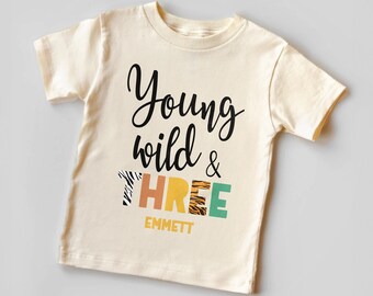 May include: A cream colored t-shirt with the text "Young wild & THREE" in black and colorful letters with animal print designs. The name "EMMETT" is printed below the text.