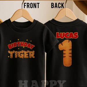 May include: Black toddler t-shirt with "BIRTHDAY TIGER" text and star graphics on the front. The back of the shirt has the name "LUCAS" and a tiger-striped number "1". The shirt is hanging on a wooden hanger.