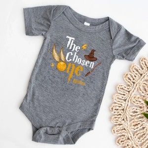 May include: Gray baby onesie with a graphic of a golden snitch, a wizard hat, and a wand. The text "The Chosen One" is printed on the onesie, with the name "Malden" printed below.