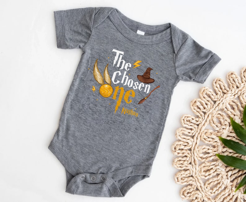 May include: Gray baby onesie with a graphic of a golden snitch, a wizard hat, and a wand. The text "The Chosen One" is printed on the onesie, with the name "Malden" printed below.