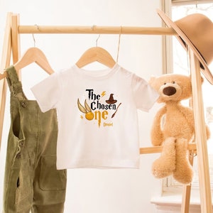 May include: White baby onesie with a gold and black graphic of a lightning bolt, a golden snitch, a wizard's hat, and a wand. The text "The Chosen One" is printed in black with the name "Daniel" printed below.