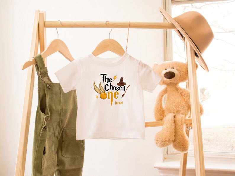 May include: White baby onesie with a gold and black graphic of a lightning bolt, a golden snitch, a wizard's hat, and a wand. The text "The Chosen One" is printed in black with the name "Daniel" printed below.