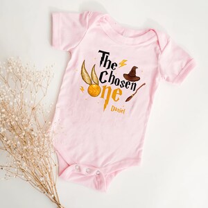 May include: Pink baby onesie with a graphic of a golden snitch, a wizard hat, and a wand. The text "The Chosen One" is printed on the onesie, with the name "Daniel" printed below.