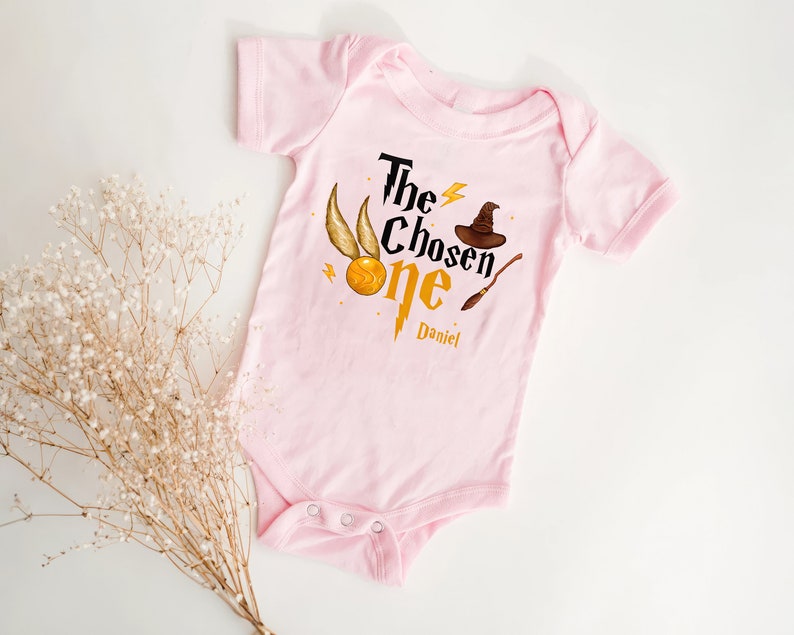 May include: Pink baby onesie with a graphic of a golden snitch, a wizard hat, and a wand. The text "The Chosen One" is printed on the onesie, with the name "Daniel" printed below.