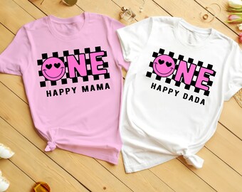 May include: Two pink and white t-shirts with a black and white checkered pattern and a pink smiley face with hearts for eyes. The text "ONE" is in pink letters and the text "HAPPY MAMA" and "HAPPY DADA" are in black letters.