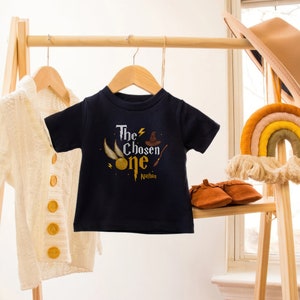 May include: A navy blue t-shirt with a graphic of a golden snitch, a wizard's hat, and a wand. The text "The Chosen One" is printed on the shirt, with the name "Nathan" underneath.