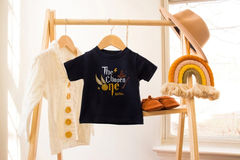 May include: A navy blue t-shirt with a graphic of a golden snitch, a wizard's hat, and a wand. The text "The Chosen One" is printed on the shirt, with the name "Nathan" underneath.