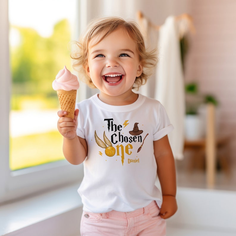 May include: A white t-shirt with a gold graphic of a lightning bolt, a golden snitch, a wizard's hat, and a wand. The text "The Chosen One" is written in black with a gold outline. The name "Daniel" is written in black below the graphic.