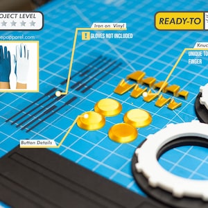 May include: A collection of gold-colored plastic pieces, including buttons and knuckle details, alongside white and black circular components. The image also features a blue cutting mat and a small image of blue and white gloves.