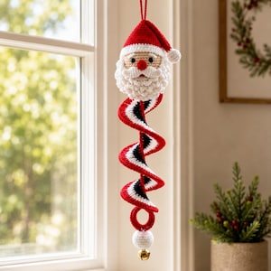 Peut inclure: Une décoration suspendue en crochet représentant le Père Noël avec un bonnet rouge, une barbe blanche et une longue queue en spirale rouge, blanche et noire. L'ornement comprend une petite sphère blanche et une clochette dorée à la base.