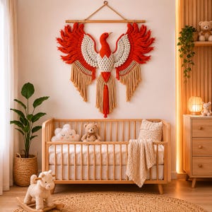 Könnte beinhalten: Ein großer Wandbehang aus Makramee in Form eines Phönix, in Rot, Weiß und Beige. Das Kunstwerk hängt über einem Holzbettchen in einem Kinderzimmer. Der Raum ist mit einem gewebten Teppich, einem Schaukelspielzeug und einer Topfpflanze dekoriert.