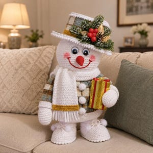 May include: A crocheted snowman figure wearing a plaid hat adorned with pine needles and red berries. The snowman has a white scarf, three white buttons, and white shoes with pom-poms. It holds a small gold gift box with red ribbon accents.
