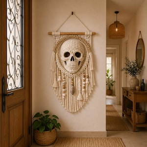 Puede incluir: Un tapiz de pared de macramé con un diseño de calavera. La calavera es de color crema y tiene cuencas de los ojos y una cavidad nasal detalladas. El macramé incluye flecos, cuentas de madera y una clavija de madera para colgar.