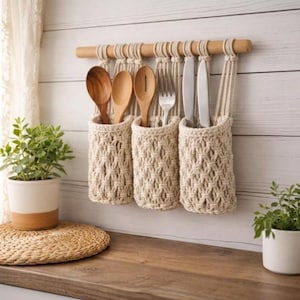 May include: A macrame utensil holder with three woven pockets suspended from a wooden dowel. The pockets hold wooden spoons, a fork, and a knife. The neutral color palette complements the natural wood and white wall.