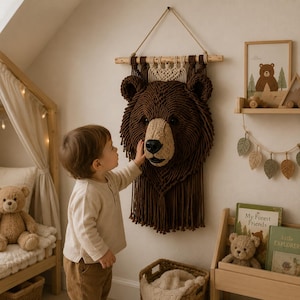 Puede incluir: Una decoración de pared con forma de cabeza de oso en macramé, hecha con cuerda de color marrón oscuro y beige. El oso presenta una melena texturizada, un hocico claro, una nariz negra y ojos oscuros, suspendido de un listón de madera horizontal.