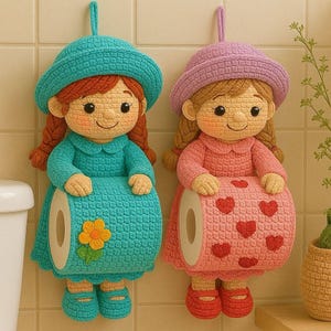 May include: Two crocheted dolls designed as toilet paper holders. The left doll wears a teal hat and dress with a yellow flower. The right doll wears a lavender hat and pink dress with red hearts. Both have braided hair and hold a roll of paper.