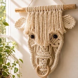 May include: Cream-colored macrame wall hanging in the shape of a bull's head. The piece features a wooden dowel at the top, dark brown button eyes, and a wooden ring for the nose. The design includes fringed details and textured patterns.