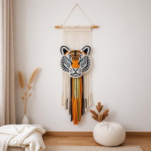 Peut inclure: Une suspension murale en macramé représentant une tête de tigre en fil orange, noir et blanc. Le visage du tigre est fixé sur une tige en bois avec de longs cordons en macramé crème, agrémentés de brins verticaux orange, verts et noirs.