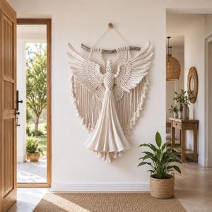 May include: A large, cream-colored macrame angel wall hanging with outstretched wings and a flowing gown. The angel is suspended from a wooden dowel and hangs against a white wall. The piece adds a touch of bohemian style to the entryway.