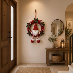 May include: A crocheted Christmas wreath featuring a central gnome with a red pointed hat, white beard, and striped legs. The wreath is decorated with green leaves, red berries, and red and white striped candy canes, hanging by a red cord.