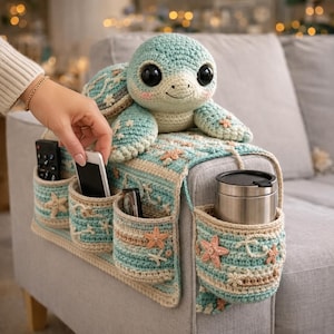 Peut inclure: Un organiseur de bras de canapé crocheté dans les tons de bleu, beige et pêche, avec des poches et une tortue de mer décorative. L'organisateur a des poches pour les télécommandes, les téléphones et un mug de voyage. La tortue de mer est bleu clair et beige.