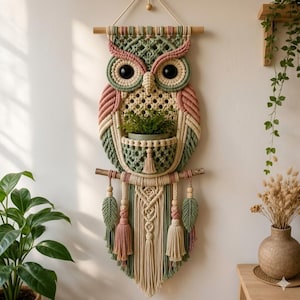 Può includere: Un arazzo in macramè a forma di gufo, realizzato con corde di cotone color verde salvia, rosa polvere e crema. Il design del gufo presenta grandi occhi neri e una tasca centrale che ospita una piccola pianta verde, con nappe e foglie.