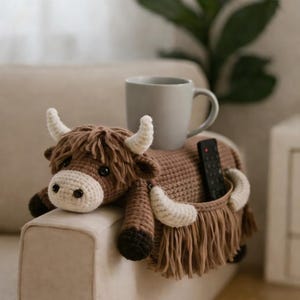 May include: A handmade crocheted Highland cow couch tidy in shades of brown, with a pocket for a remote control. The cow has white horns and a white muzzle. A grey mug rests on the caddy.