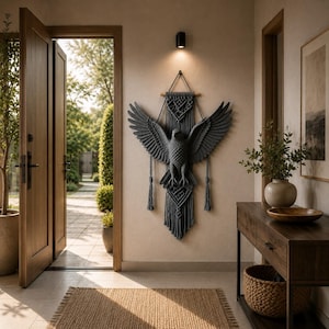May include: A dark gray macrame wall hanging featuring a central bird with outstretched wings, suspended from a wooden dowel. The intricate knotted design includes geometric patterns and long, dangling fringe tassels at the bottom edges.