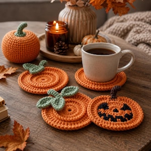 Peut inclure: Un ensemble de dessous de verre en crochet en forme de citrouille orange, dont un avec un visage de jack-o'-lantern noir et d'autres avec des feuilles vertes. Une tasse en céramique beige repose sur un dessous de verre, près d'une citrouille.