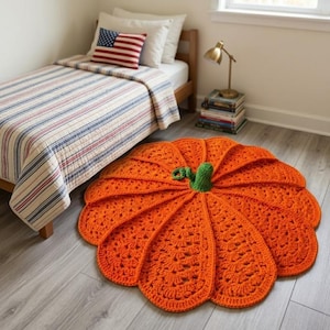 May include: A round, bright orange crochet rug shaped like a pumpkin, featuring textured, fan-like segments radiating from a central green stem. The rug is placed on a light-colored wooden floor in a room with a bed and a small lamp.