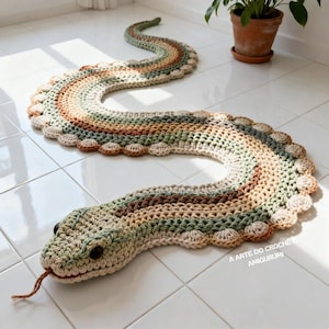 Könnte beinhalten: Ein gehäkelter Schlangen-Teppich in Grün-, Braun- und Beigetönen. Der Teppich hat einen detaillierten Kopf und einen langen, gewundenen Körper. Der Text "A ARTE DO CROCHÊ & AMIGURUMI" ist sichtbar. Der Teppich liegt auf einem gefliesten Boden.