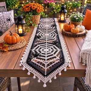Peut inclure: Un chemin de table rectangulaire en crochet présentant un motif de toile d'araignée noir et blanc avec de petites araignées noires et des pompons décoratifs sur les bords, posé sur une table en bois avec des citrouilles et des lanternes.