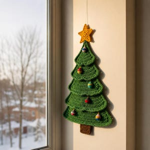 Puede incluir: Un adorno de pared en forma de árbol de Navidad tejido a ganchillo en color verde, con una estrella amarilla en la punta y un tronco marrón en la base. El árbol está decorado con pequeños adornos tejidos de colores y diversos patrones.