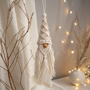 May include: A handmade macrame gnome ornament with a cream-colored pointed hat and long, flowing beard. The gnome has a wooden nose and is suspended by a cream-colored string. The background includes soft lighting and decorative elements.