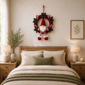 May include: A circular red crocheted wreath featuring a central gnome with a tall red hat, a white beard, and striped legs. The wreath is decorated with green fabric leaves, red berries, and small red and white striped candy cane accents.