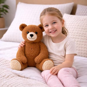 Puede incluir: Un oso de peluche de ganchillo marrón hecho a mano con hocico y almohadillas de los pies color crema. El oso tiene una nariz bordada en negro. El oso mide aproximadamente 35 cm de alto y está sentado en una cama.
