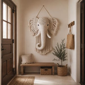 Peut inclure: Décoration murale en forme de tête d'éléphant en macramé crème, avec des oreilles frangées et une longue trompe. L'éléphant est accroché à un mur crème. La pièce comprend également un banc en bois, un panier tressé et une petite plante.