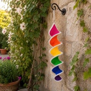 Peut inclure: Une décoration suspendue en crochet en forme de spirale présentant un dégradé de couleurs arc-en-ciel vif composé de segments rose, orange, jaune, vert, bleu et violet, chacun bordé d'une bordure blanche, suspendue par un cordon blanc et un anneau.