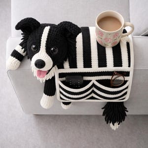 Puede incluir: Un organizador de reposabrazos de sof&aacute; tejido en crochet blanco y negro con dise&ntilde;o de perro, lengua rosa, dos bolsillos frontales con un mando a distancia negro y gafas de montura marr&oacute;n, y una taza de cer&aacute;mica floral en la parte superior.