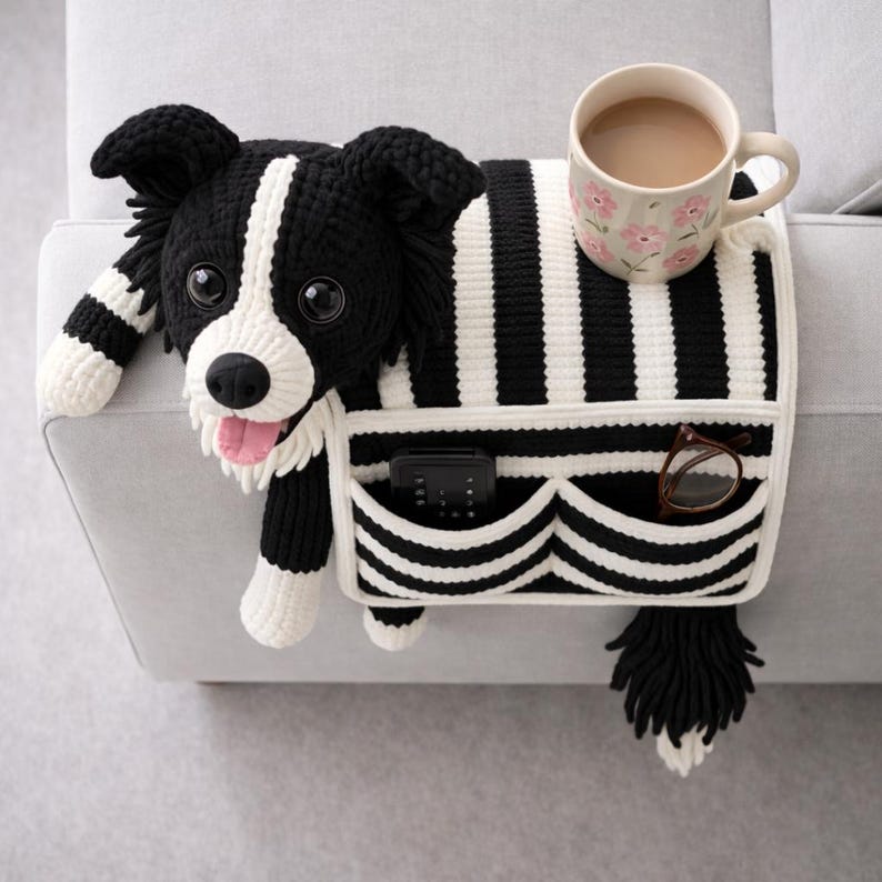 Puede incluir: Un organizador de reposabrazos de sof&aacute; tejido en crochet blanco y negro con dise&ntilde;o de perro, lengua rosa, dos bolsillos frontales con un mando a distancia negro y gafas de montura marr&oacute;n, y una taza de cer&aacute;mica floral en la parte superior.