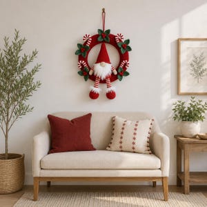 May include: A crocheted Christmas wreath featuring a central gnome with a red hat, white beard, and striped legs. The wreath is decorated with green leaves, red berries, and red and white candy canes, all suspended by a red loop.