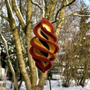 May include: A handcrafted, spiral-shaped crochet decoration in shades of red, yellow, and black, suspended from a white string. The ornament hangs against a blurred winter backdrop of bare trees and snow.
