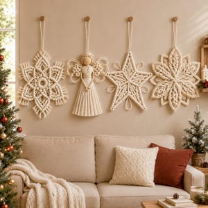 Op de afbeelding: Vier off-white macramé wandhangers die aan de muur hangen. De set bevat een grote sneeuwvlok, een centrale engelfiguur met een houten kraal als hoofd, een vijfpuntige ster en een complex geometrisch bloemvormig ornament van katoenen koord.